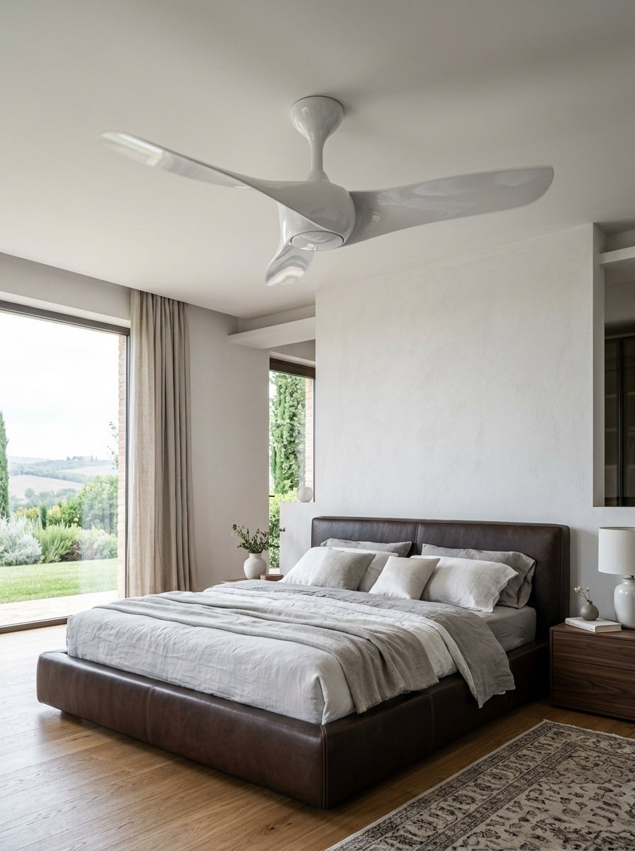 A sleek white ceiling fan with three curved blades hanging over a leather bed in a bright, modern master bedroom.