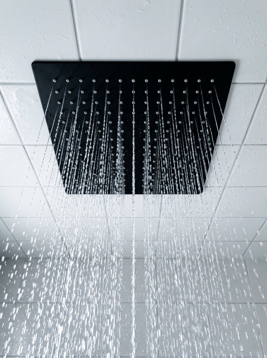 A matte black square shower head mounted on a white tiled ceiling, pouring streams of water downwards.