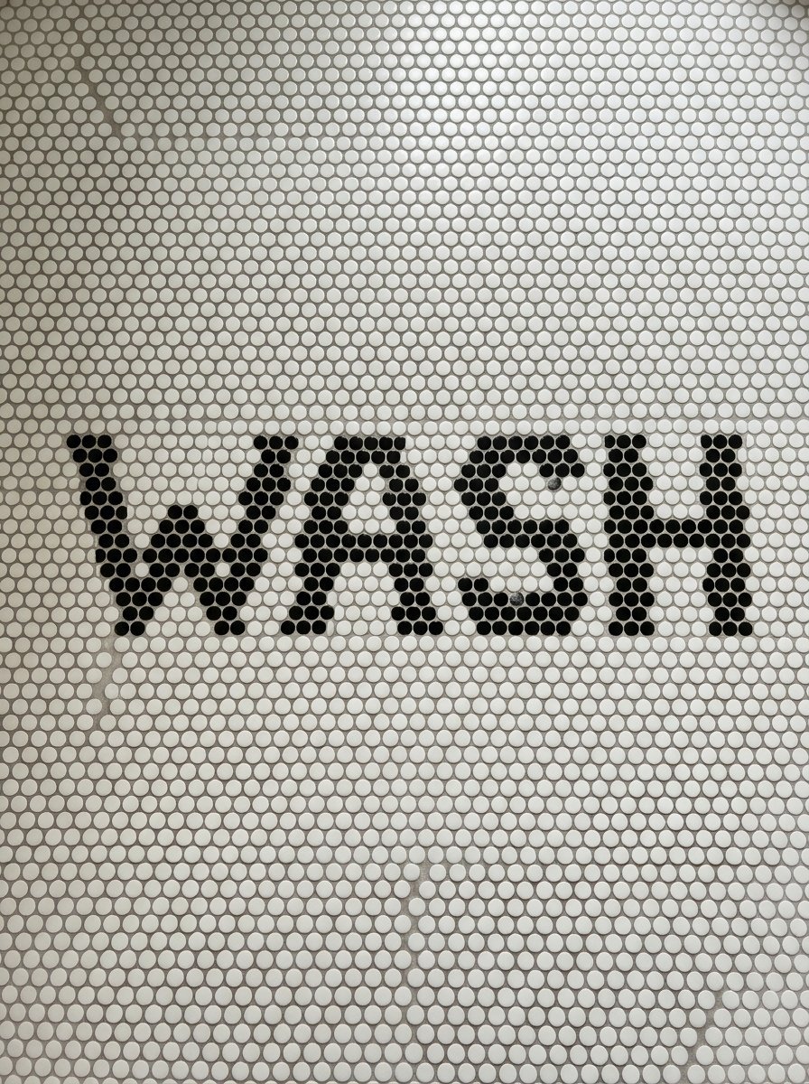 The word Wash spelled out in bold black penny tiles against a field of white circular bathroom floor tiles.